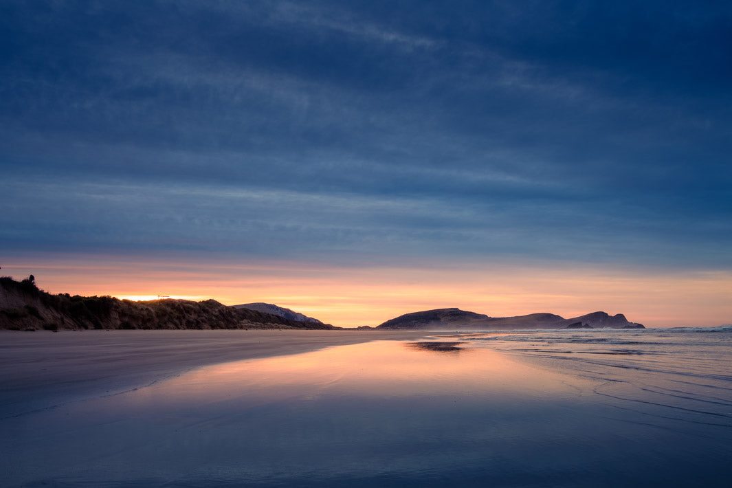 Colourful stripes of blue and orange in the sky at sunrise over a bay of water and sand.