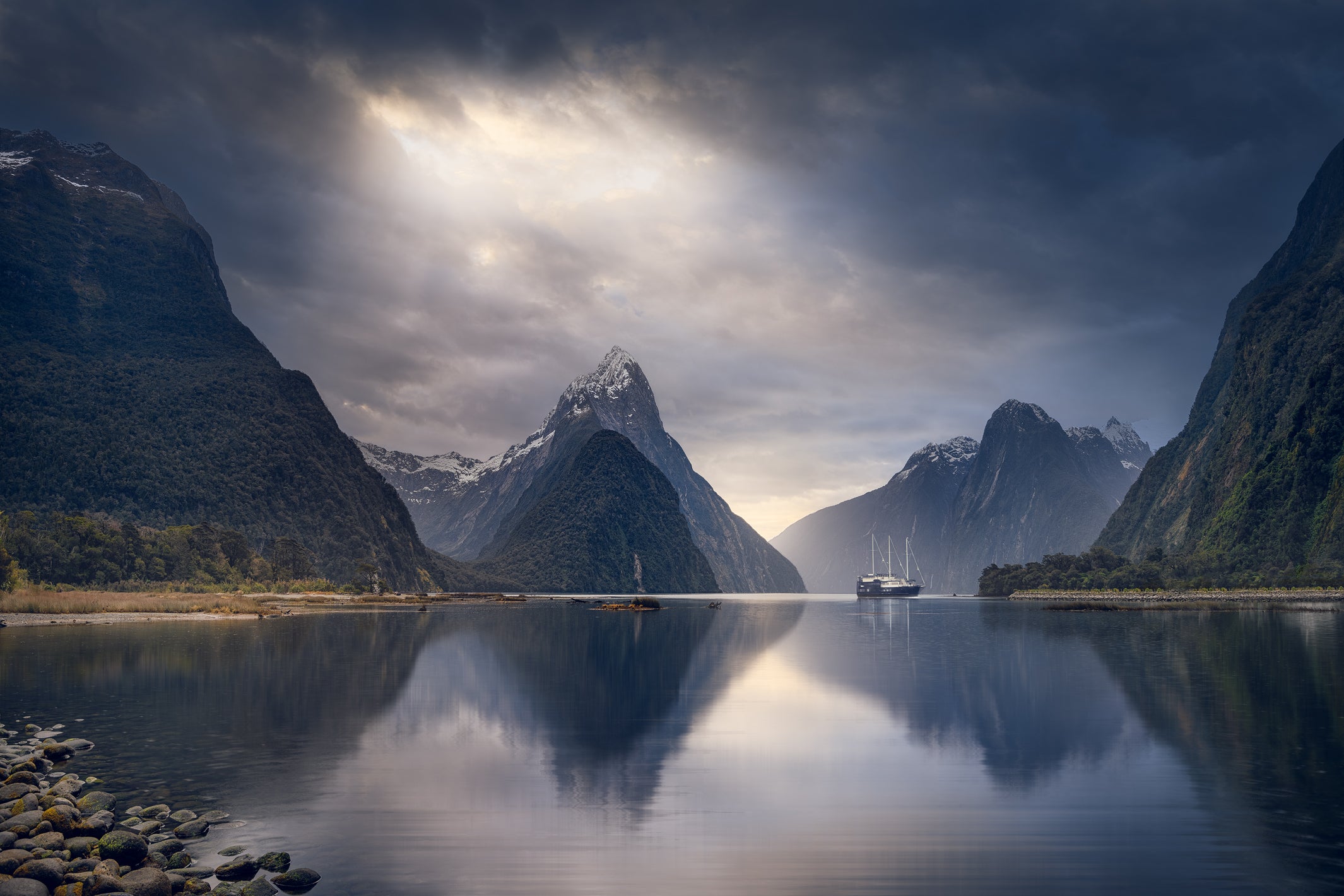 NZ Landscape photo print of mountains in Milford Sound, with moody dark skies above and still reflective water below. There is a boat in the background.