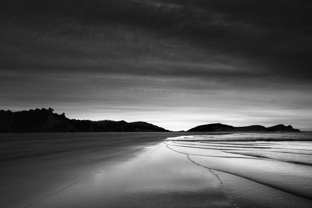 Black and white photo of wave lines leading into the centre with hills in the background and dark clouds above