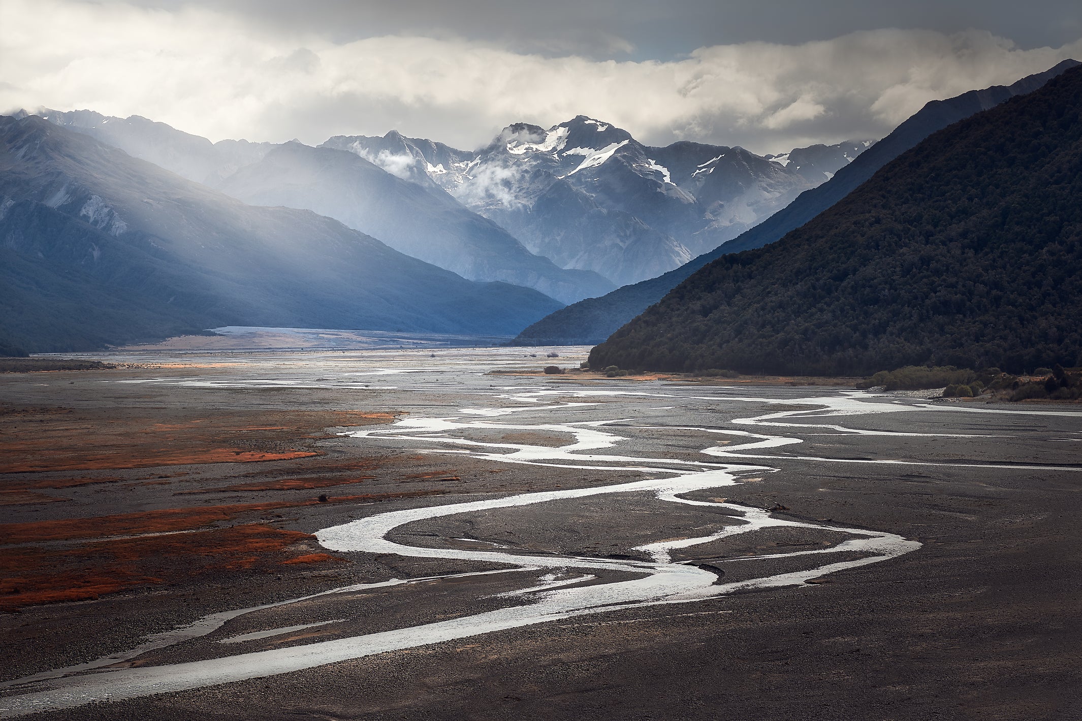 snow capped mountains with clouds above and sunrays over the valley & river below 