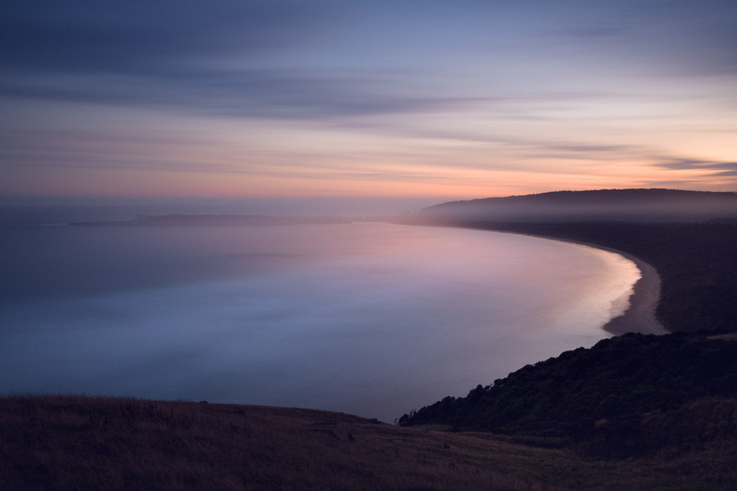 View out from trees and hills down to the sea at sunset, with pink and blue colours in the sky.