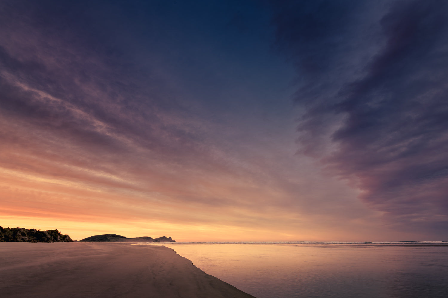 Dramatic dark clouds lit by the yellow of sunrise, above a bay of water.