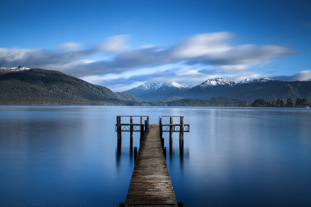 still blue lake with a wooden jetty in the centre and snow topped mountains in the background