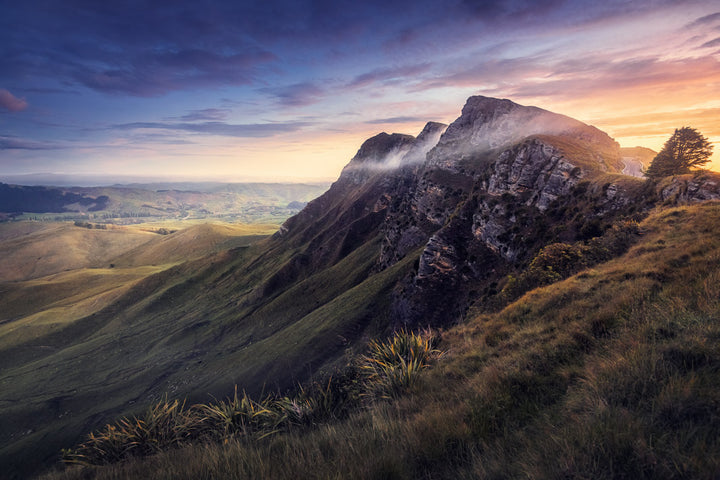 NZ landscape photo print of sunset at the top of a hill overlooking hills and fields. There is a wisp of cloud at the top of the peak.