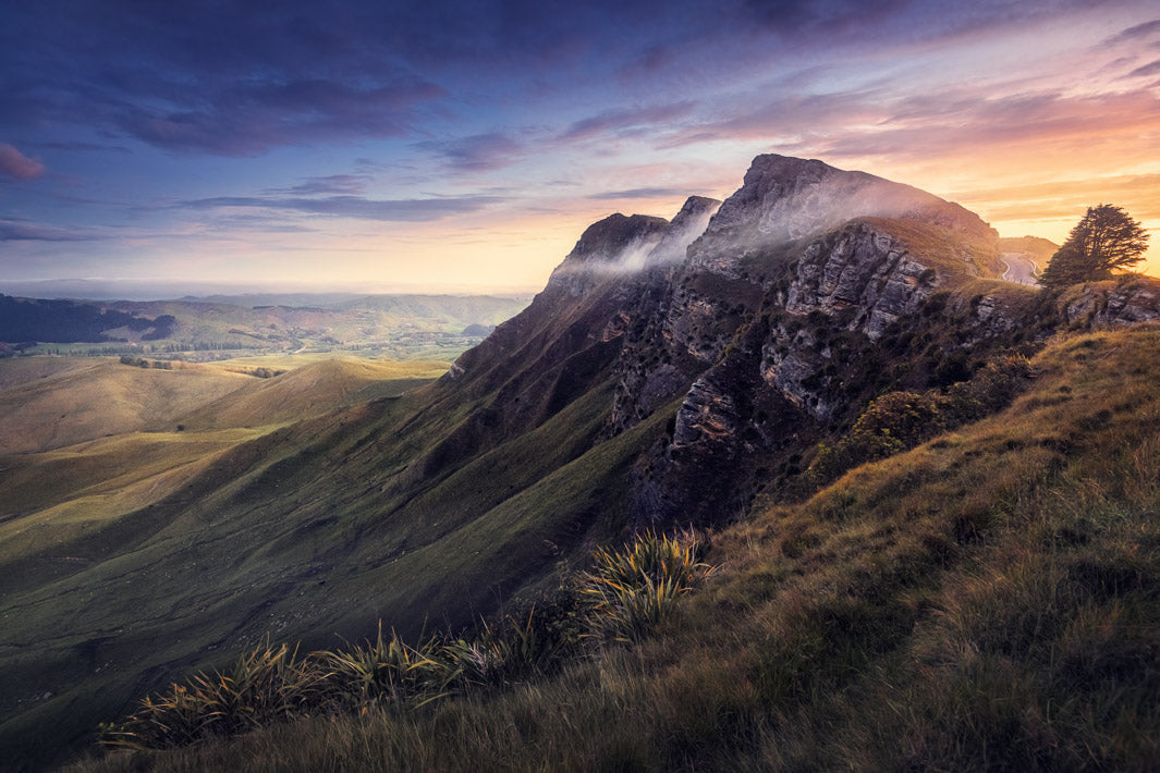 landscape photo of the sun setting behind high mountain peaks with grasses in the foreground and hills in the background