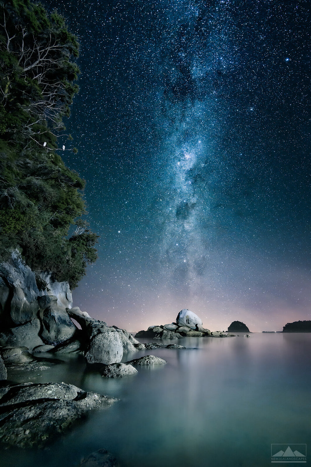 Vertical photo from New Zealand of the Milky Way above rocks and the sea, taken at night with pink and blue pastel colours.